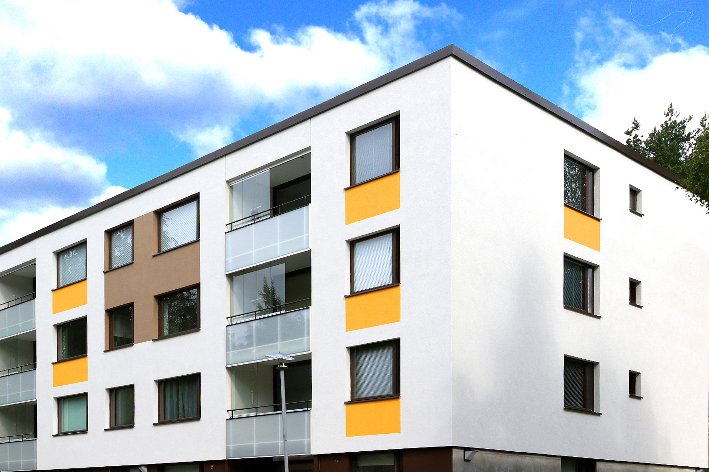 Valkoinen kolmikerroksinen asuintalo, jossa ikkunoiden välejä korostettu keltaisella ja ruskealla.
